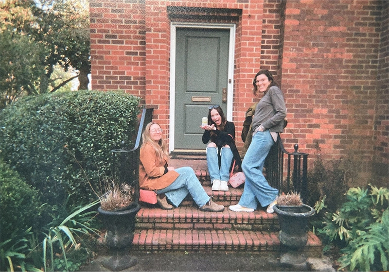 Abby, Morgan and Jenny sitting on brick steps in front of a house with a green door, surrounded by greenery.